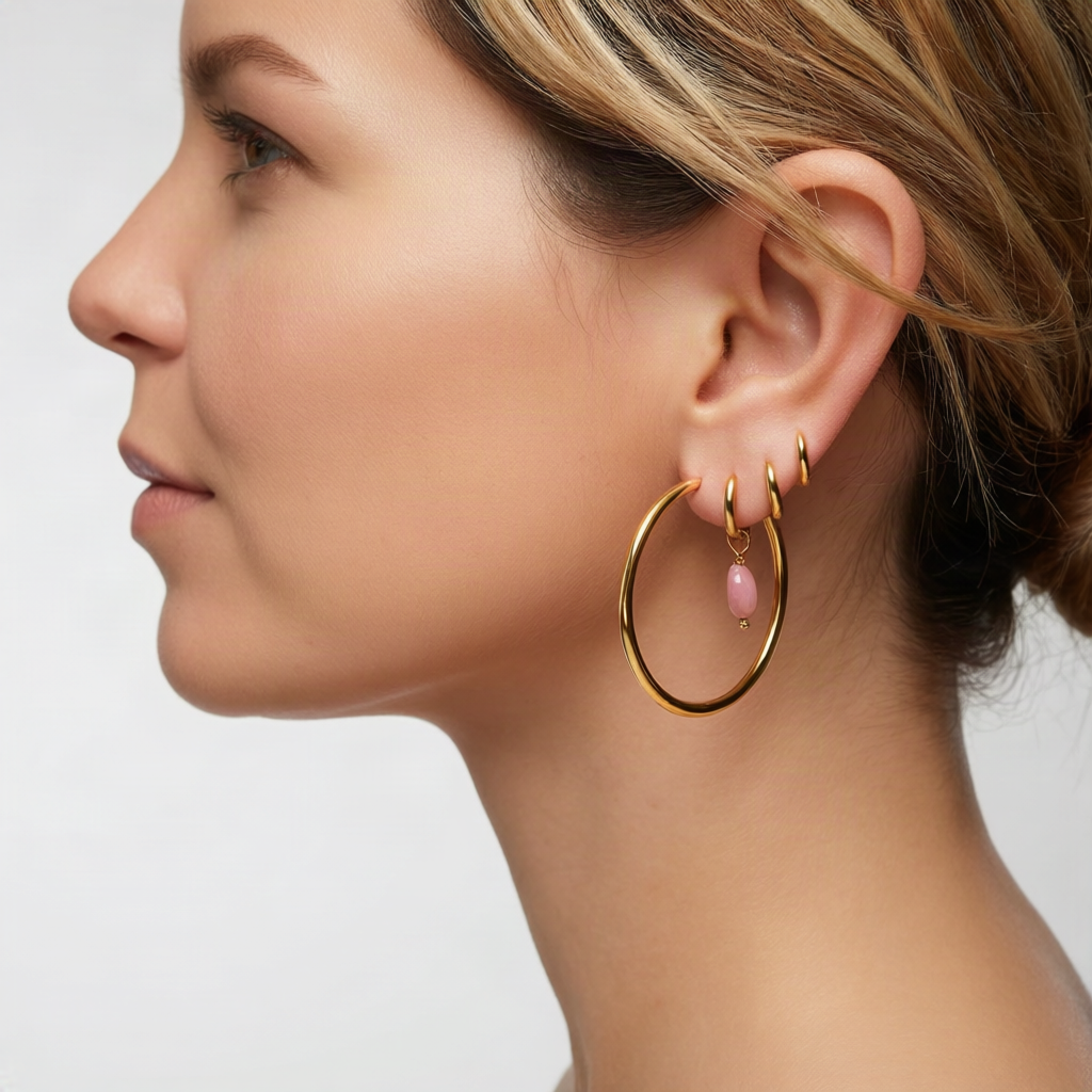 A close-up side profile of a woman wearing a pair of gold hoop earrings with a small dangling pink bead, shown against a clean light gray background.