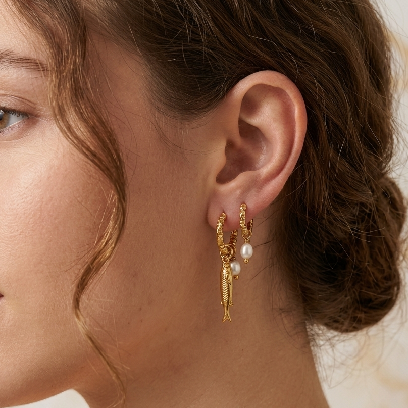 A close-up side profile of a woman wearing a delicate gold hoop earring with a dangling gold leaf charm and small white pearl accents.