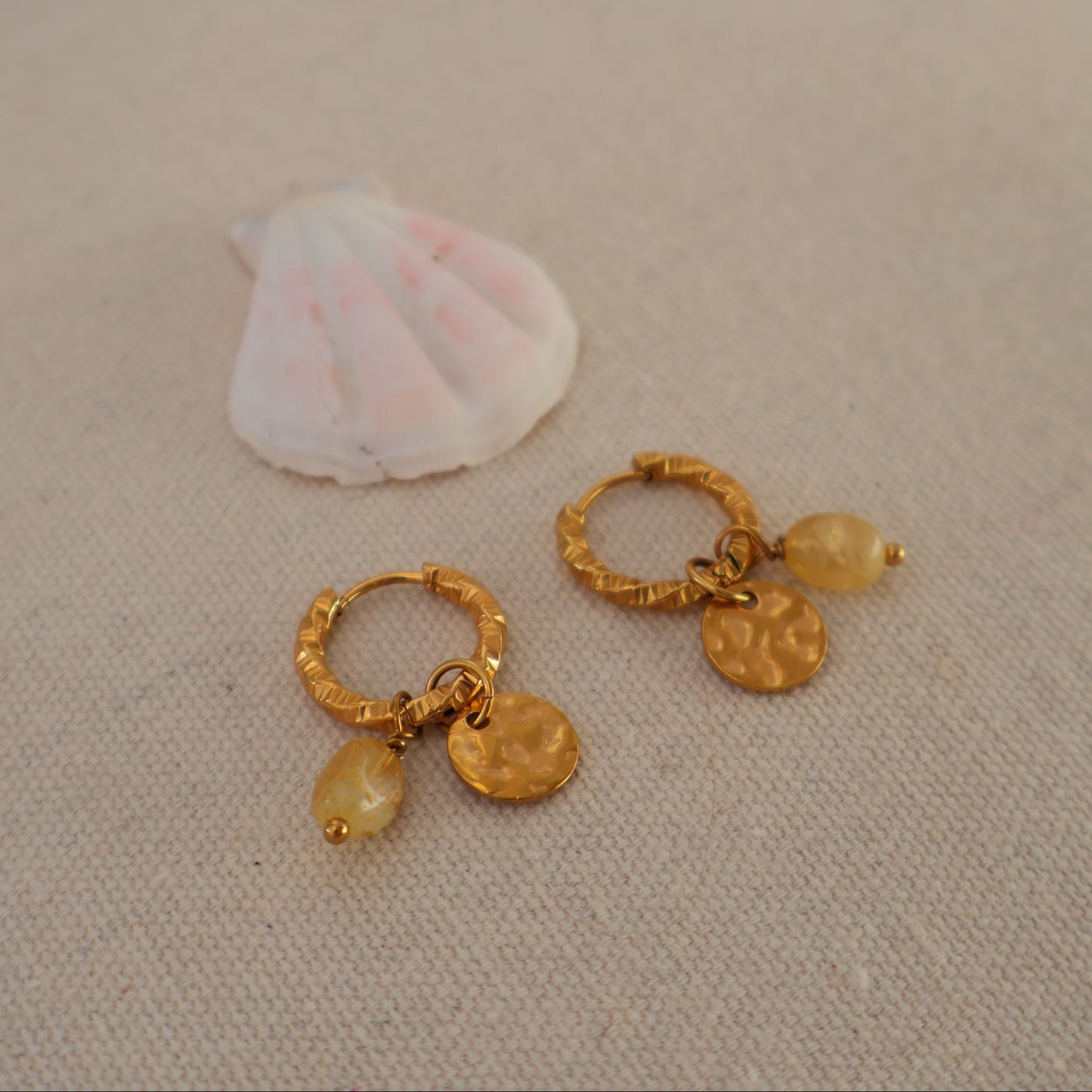 A pair of gold hoop earrings with small dangling charms, displayed on a beige fabric surface next to a white seashell.