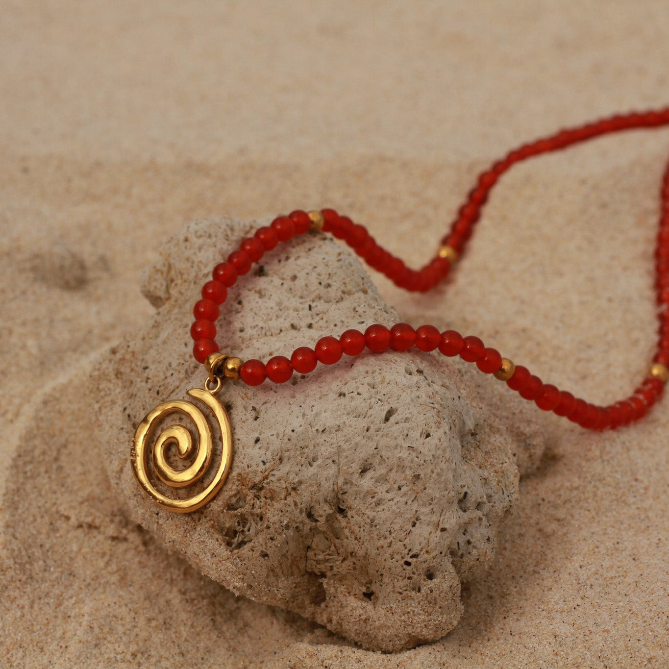 Red carnelian spiral necklace with a gold swirl pendant resting on a sandy surface.