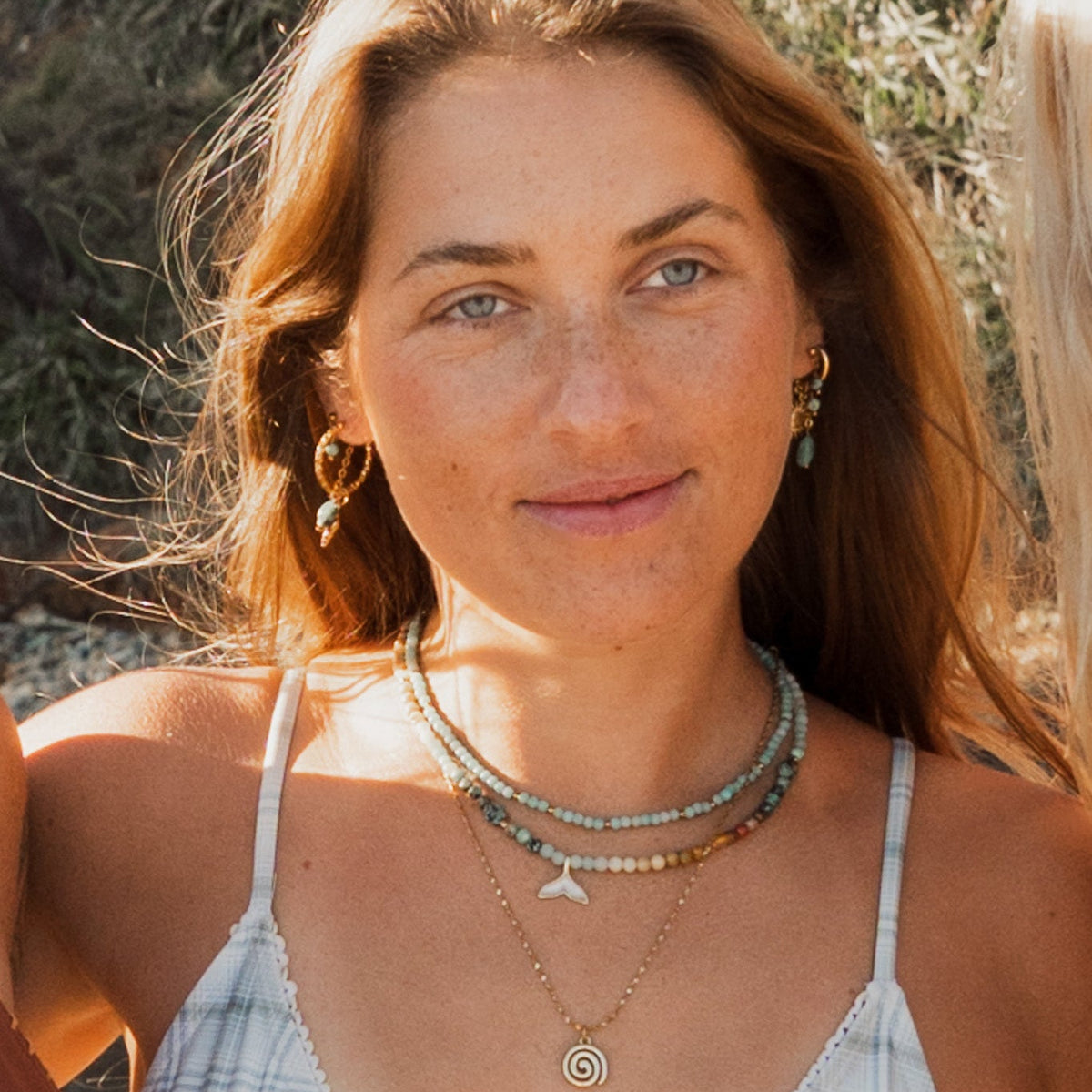 A close-up lifestyle shot of a woman outdoors wearing layered gold necklaces, including a spiral pendant, a whale tail charm, and a beaded strand, along with a white and blue plaid bikini top.