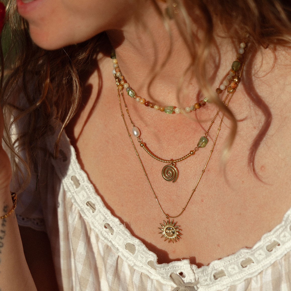 A close-up lifestyle shot of a woman wearing a layered bohemian-style gold necklace set. The set includes a main gold chain with a sun pendant, a middle chain with a spiral pendant and pearl accent, and a shorter beaded chain.