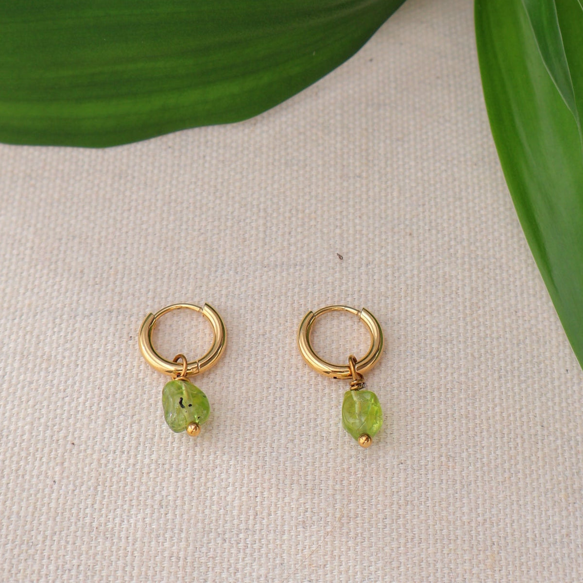 Gold earrings with green stones on a beige surface with green leaves.
