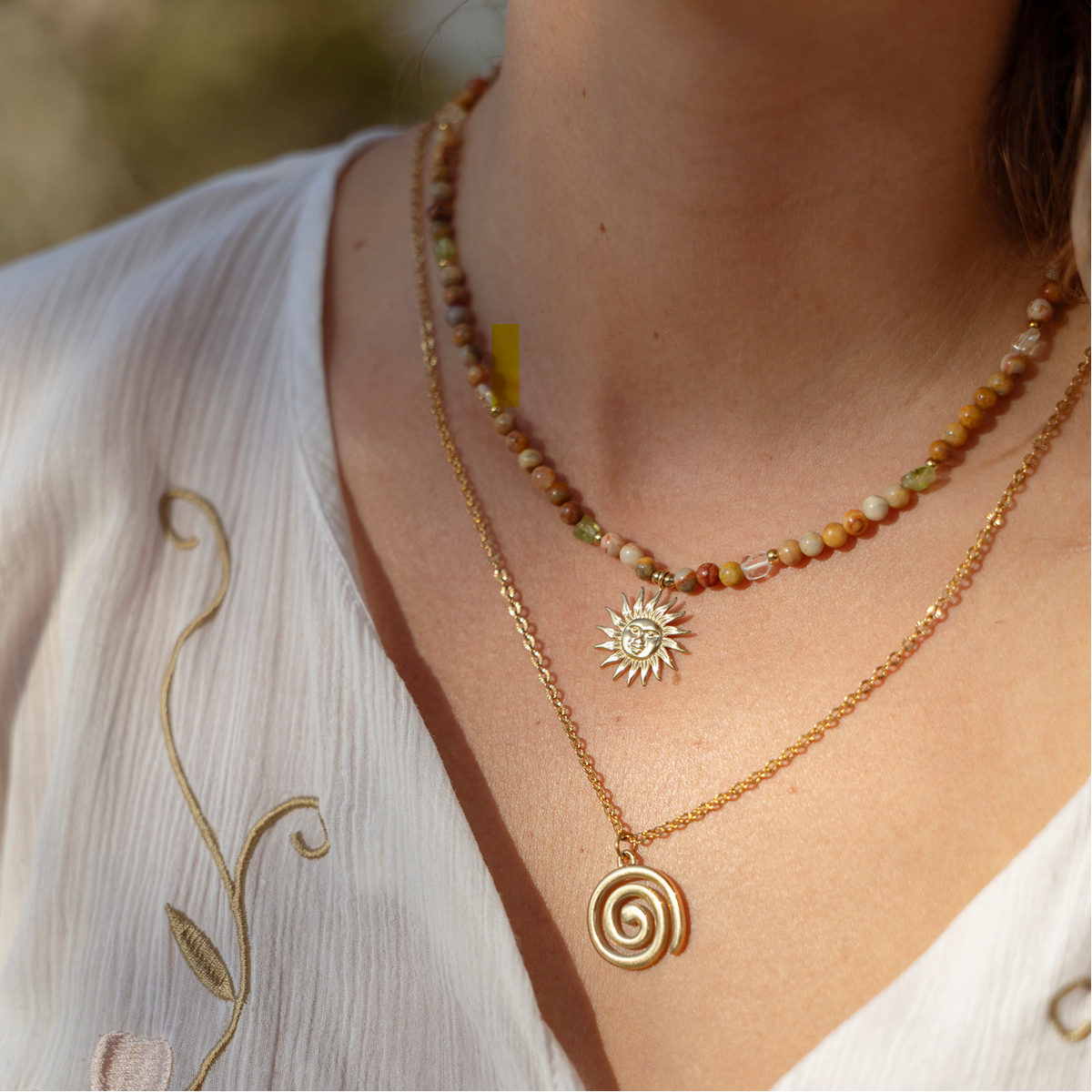 A close-up of a person wearing layered necklaces: a gold chain necklace with a spiral sun pendant and a shorter beaded necklace with earthy beads and a small central stone charm.