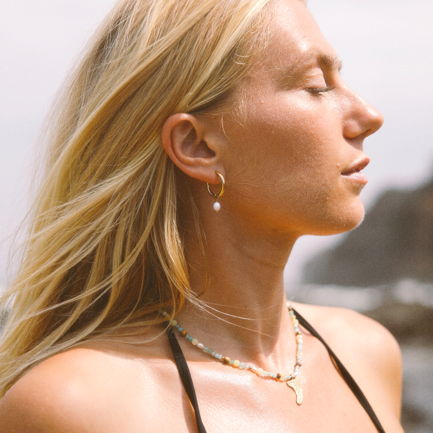 A close-up of a person wearing a beaded necklace with a gold spiral pendant. The necklace features small multicolored beads in pastel shades of orange, teal, light blue, and white.