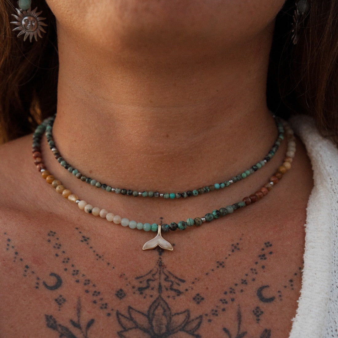 A beaded necklace featuring a gold coin pendant with a figure, accented by light green, turquoise, and orange marbled beads on a textured beige fabric background.