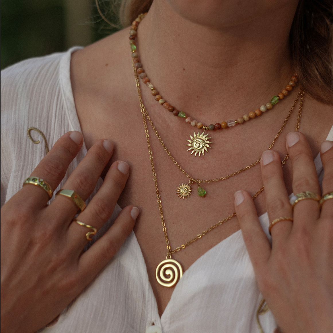 A close-up of a person wearing layered gold necklaces and rings, with their hands touching their chest. The necklaces include a beaded choker, a sun pendant necklace, a spiral pendant necklace, and a delicate chain with a small sun and green bead detail.