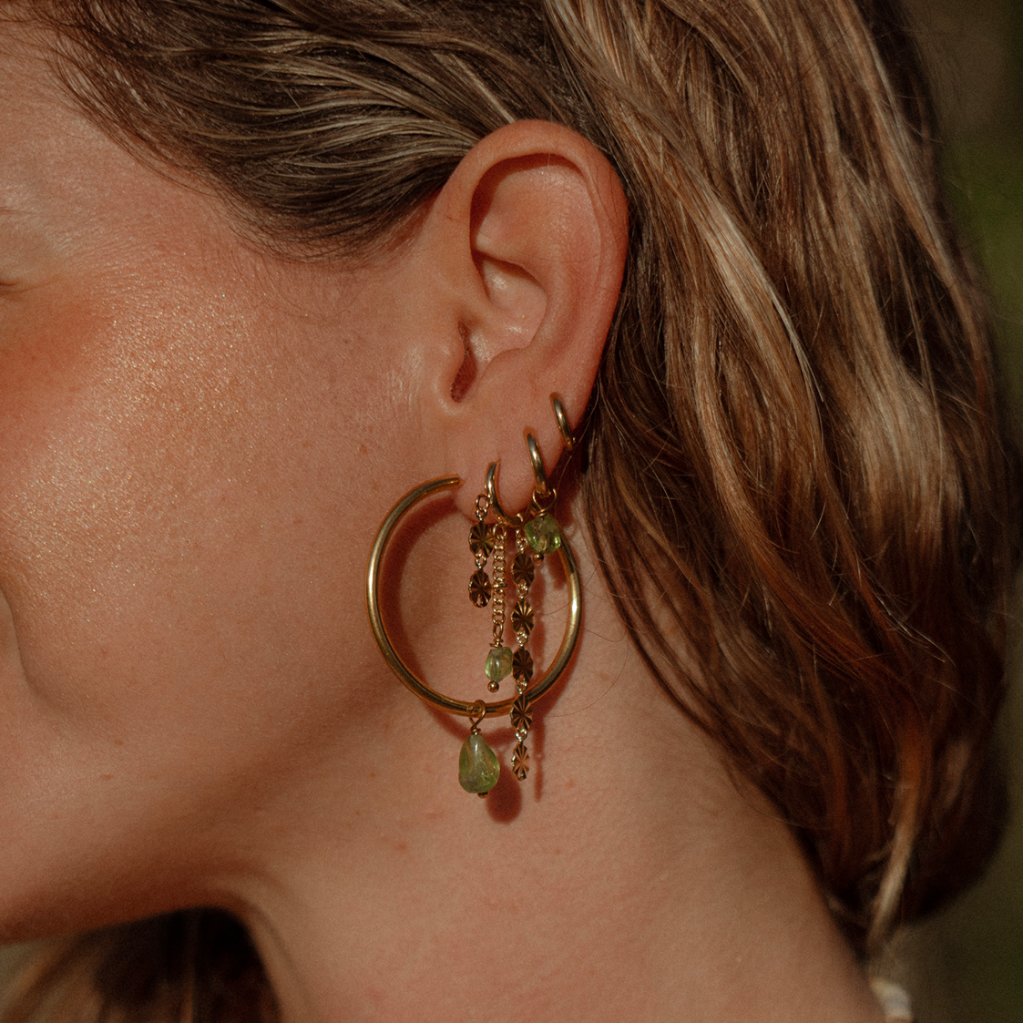 A woman in profile on a beach at sunset, wearing silver hoop earrings with dangling sunburst charms, a beaded necklace, and a white towel over her shoulder.
