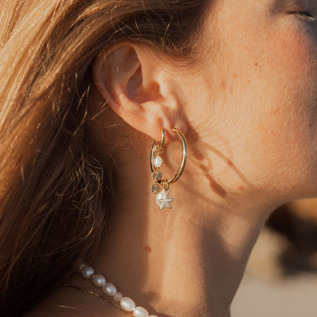 A close-up side profile of a woman wearing a gold hoop earring with a dangling white pearl star charm, along with a pearl necklace, in warm golden-hour sunlight.