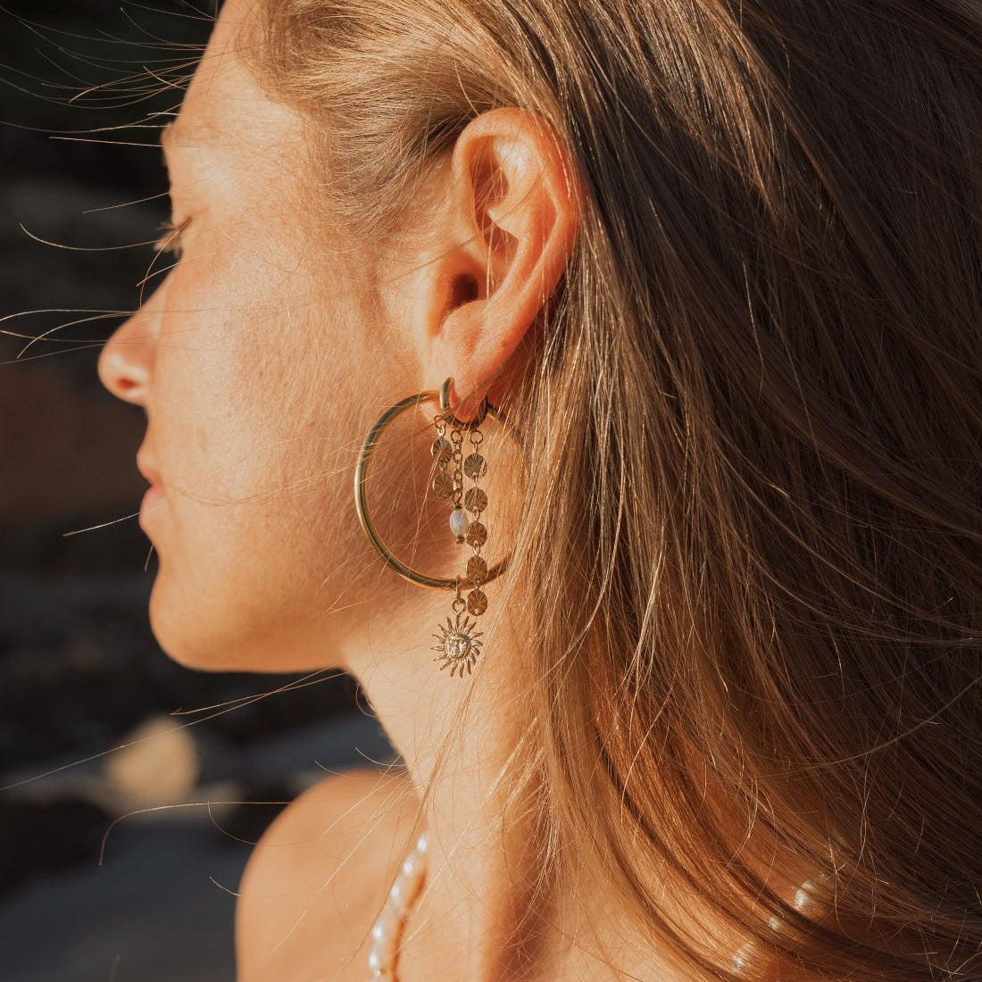 A close-up side profile of a woman outdoors wearing a large gold hoop earring with dangling sunburst and dot charms, along with layered pearl and gold necklaces.
