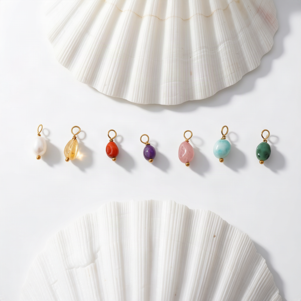 Nine small gemstone jewelry charms or swatches in assorted colors are arranged in a row on a white surface, with two large white seashell props above and below the items.