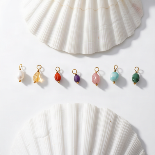 Nine small gemstone jewelry charms or swatches in assorted colors are arranged in a row on a white surface, with two large white seashell props above and below the items.