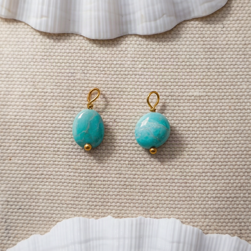 A pair of gold drop earrings with oval turquoise-blue stone pendants, displayed on a textured beige fabric background with white seashell props at the top and bottom.