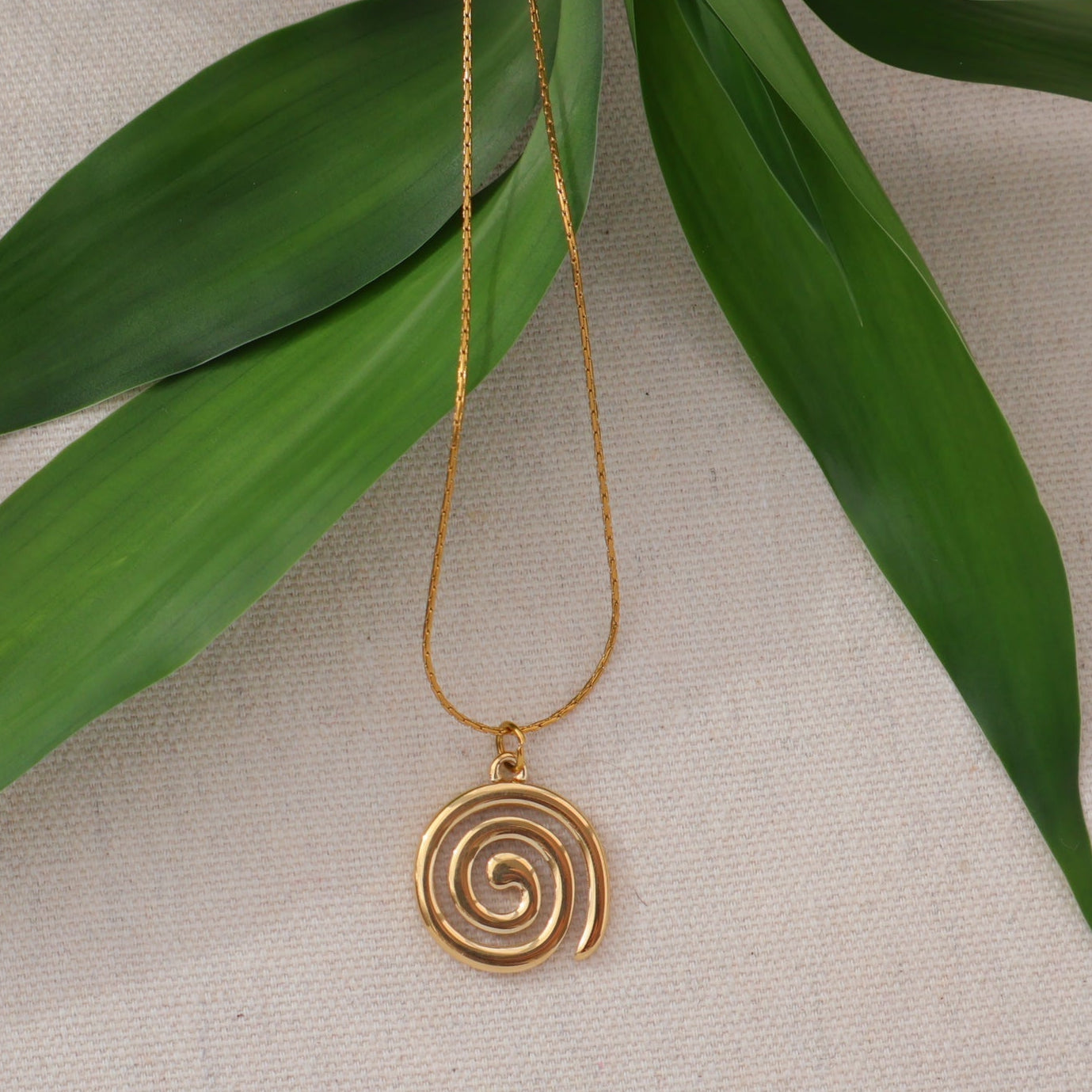 Gold spiral pendant necklace on a beige background with green leaves