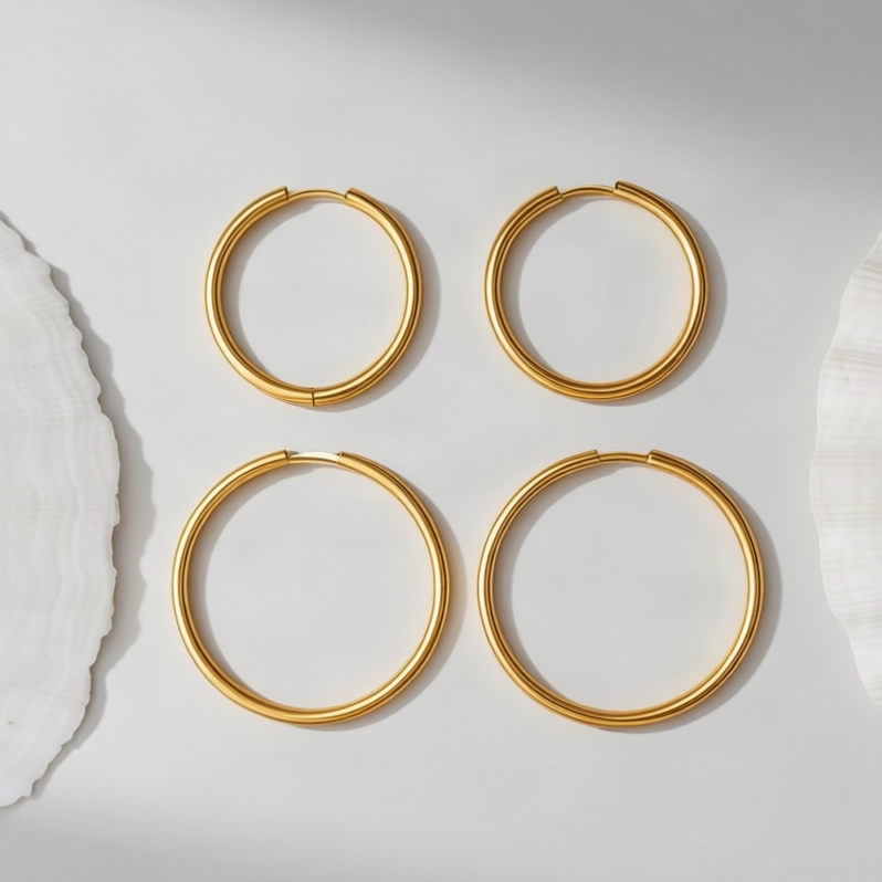 Four gold hoop earrings arranged in a 2x2 grid on a light gray surface, flanked by white seashell props.