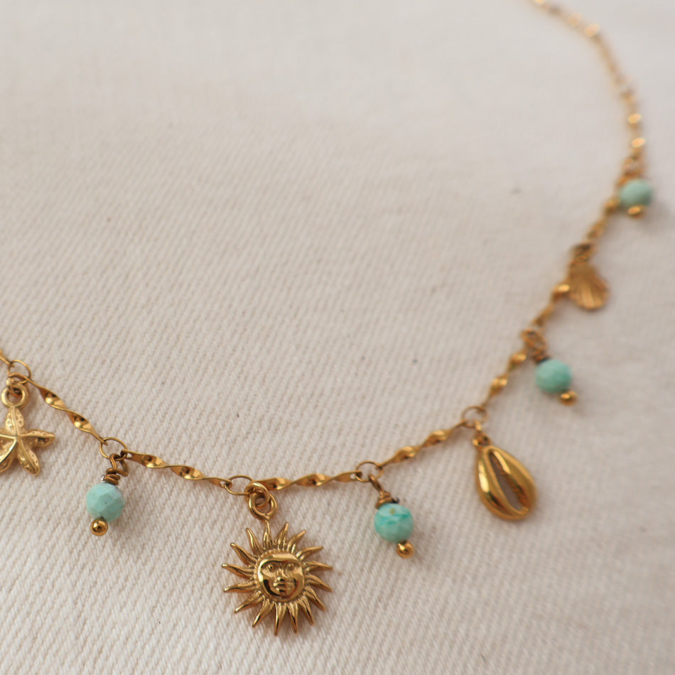 A gold-toned beaded necklace with various shell and sun pendant charms, featuring light blue turquoise beads, displayed on a textured off-white fabric background.