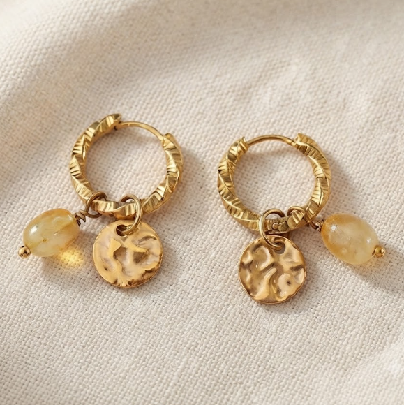 A pair of gold hoop earrings with textured circular charms and small oval bead drops, displayed on a light beige fabric background.