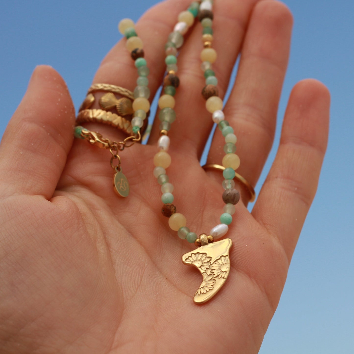 Hand holding a beaded necklace with a gold pendant against a clear blue sky