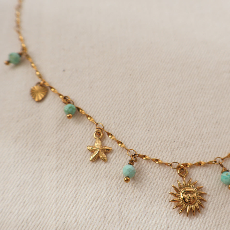 A gold chain necklace with small dangling charms, including a sun face, starfish, shell, and turquoise beads, laid on a textured off-white fabric background.