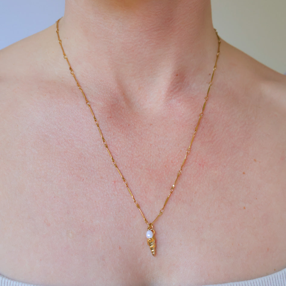 A delicate gold chain necklace with a small spiral shell pendant and a white pearl, shown worn on a model's neck.