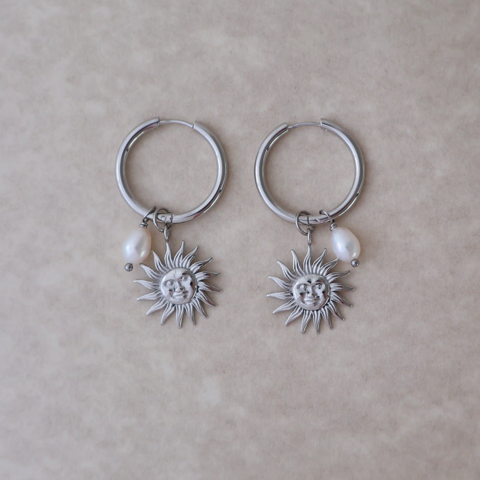 A pair of silver hoop earrings with dangling sun face charms and small white pearl beads, displayed on a neutral beige textured background.