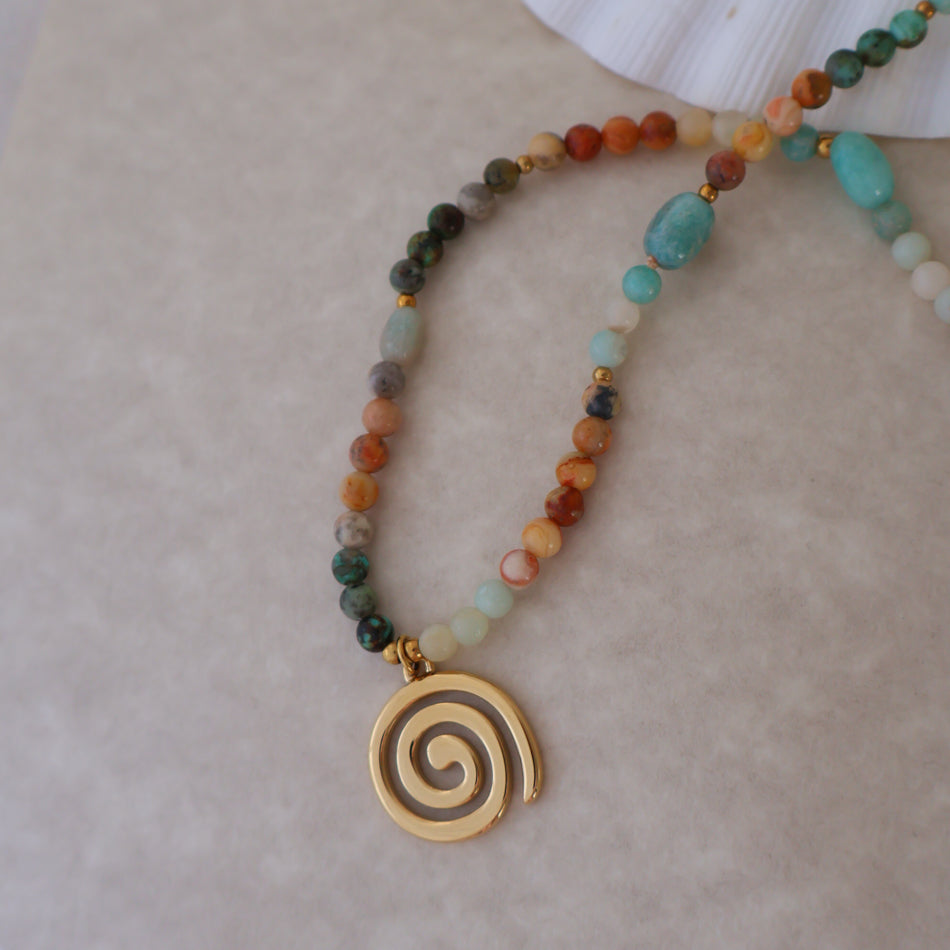 A beaded necklace with a gold spiral pendant, featuring multi-colored stone beads in earthy tones and turquoise accents, displayed on a neutral textured surface.