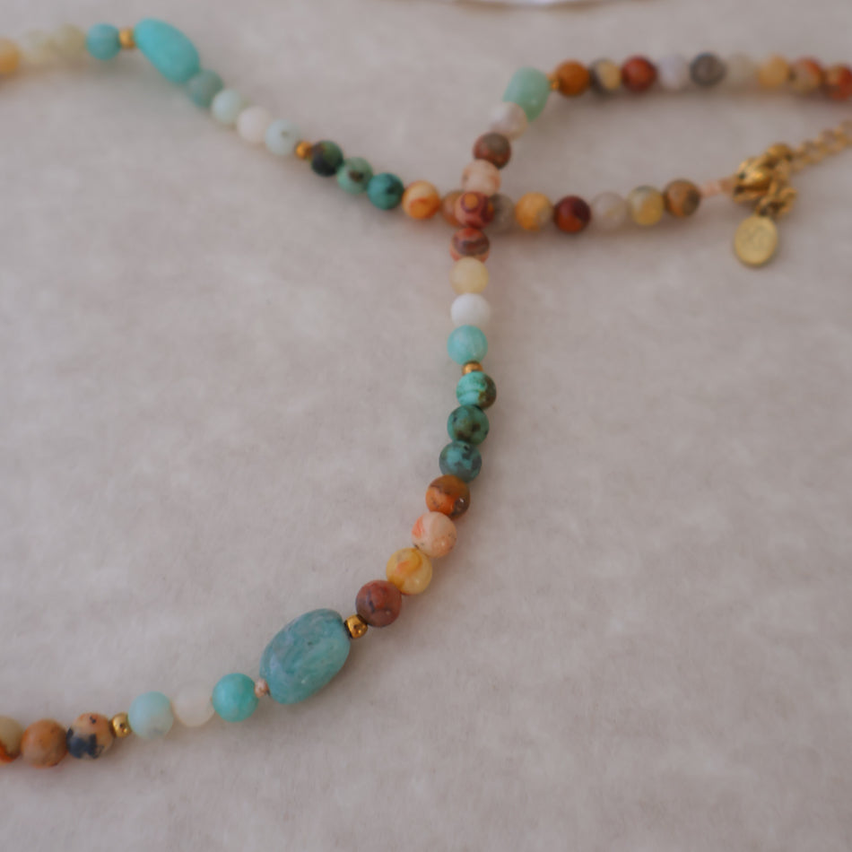 A beaded necklace with a mix of turquoise, brown, orange, white, and green stones, featuring a large turquoise stone and a gold-tone clasp, displayed on a light beige textured surface.