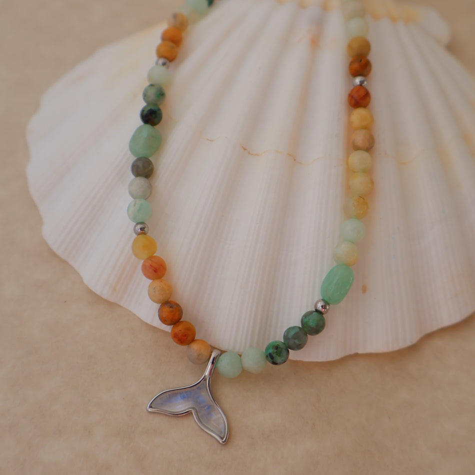 A beaded necklace with a silver whale tail pendant, displayed on a white seashell against a beige background. The beads feature a mix of earthy orange, sage green, yellow, and dark gray stone.