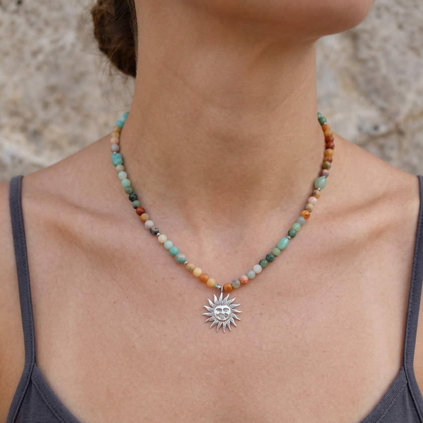 A close-up of a woman wearing a beaded necklace with a silver sun pendant. The necklace features multi-colored beads in shades of turquoise, orange, brown, and green, with a silver sun charm at the center.