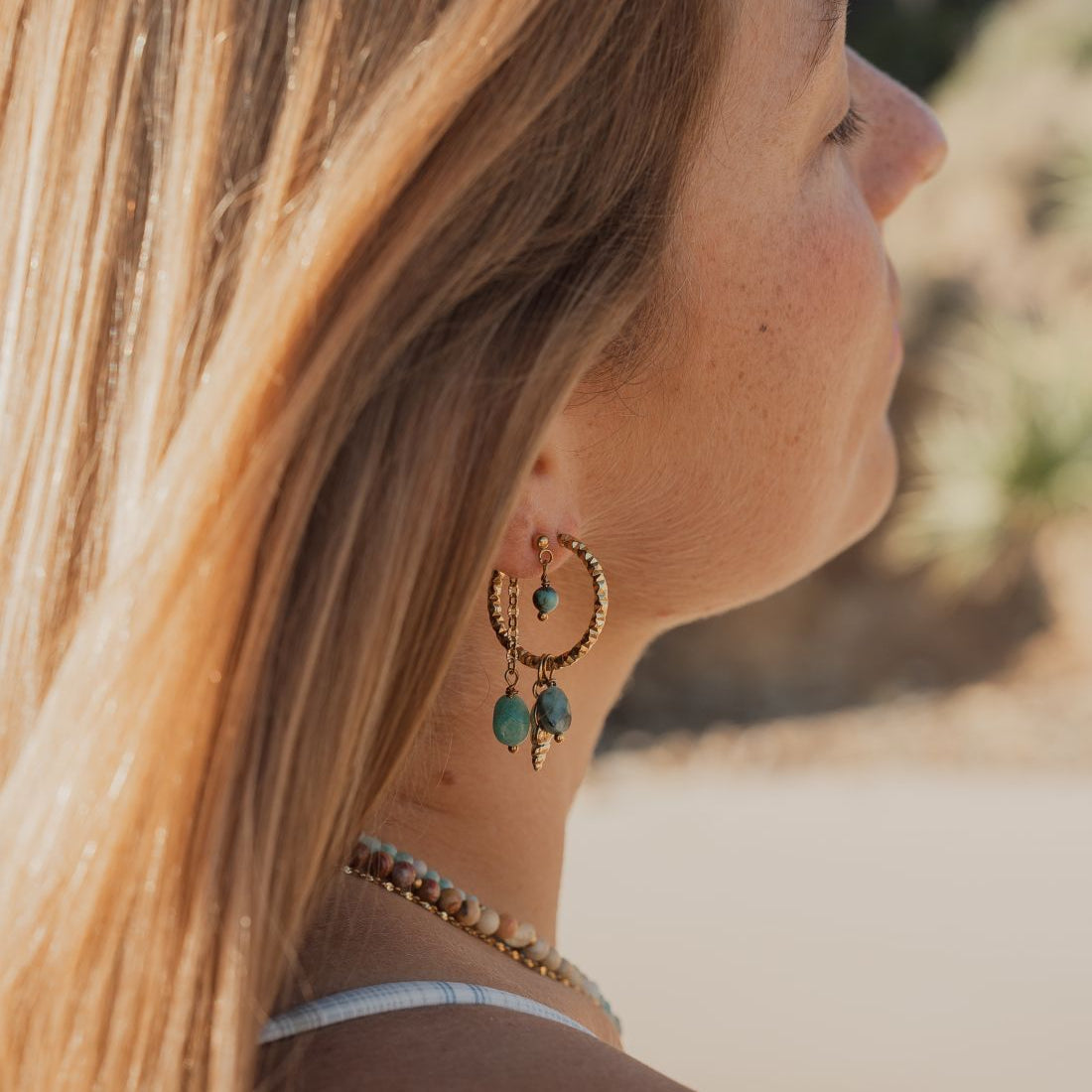 A close-up side profile of a woman with long blonde hair wearing a gold crescent hoop earring with dangling turquoise green beads and a matching beaded necklace.