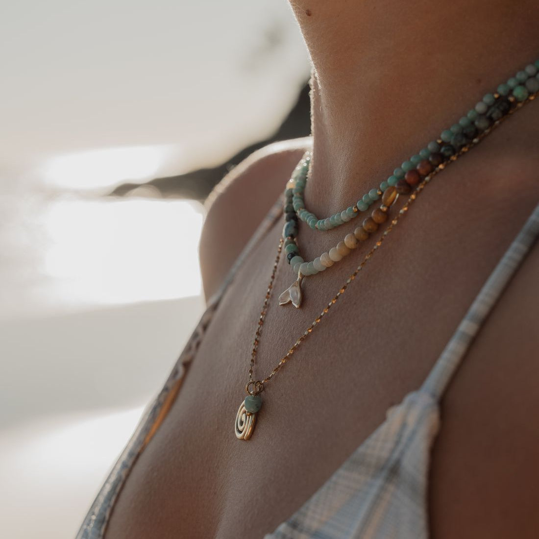 A close-up lifestyle shot of a woman's neck and chest wearing a layered necklace set. The set includes a gold chain with a small turquoise and white bead pendant, a gold chain with a small white shell charm, and a turquoise and white beaded strand.
