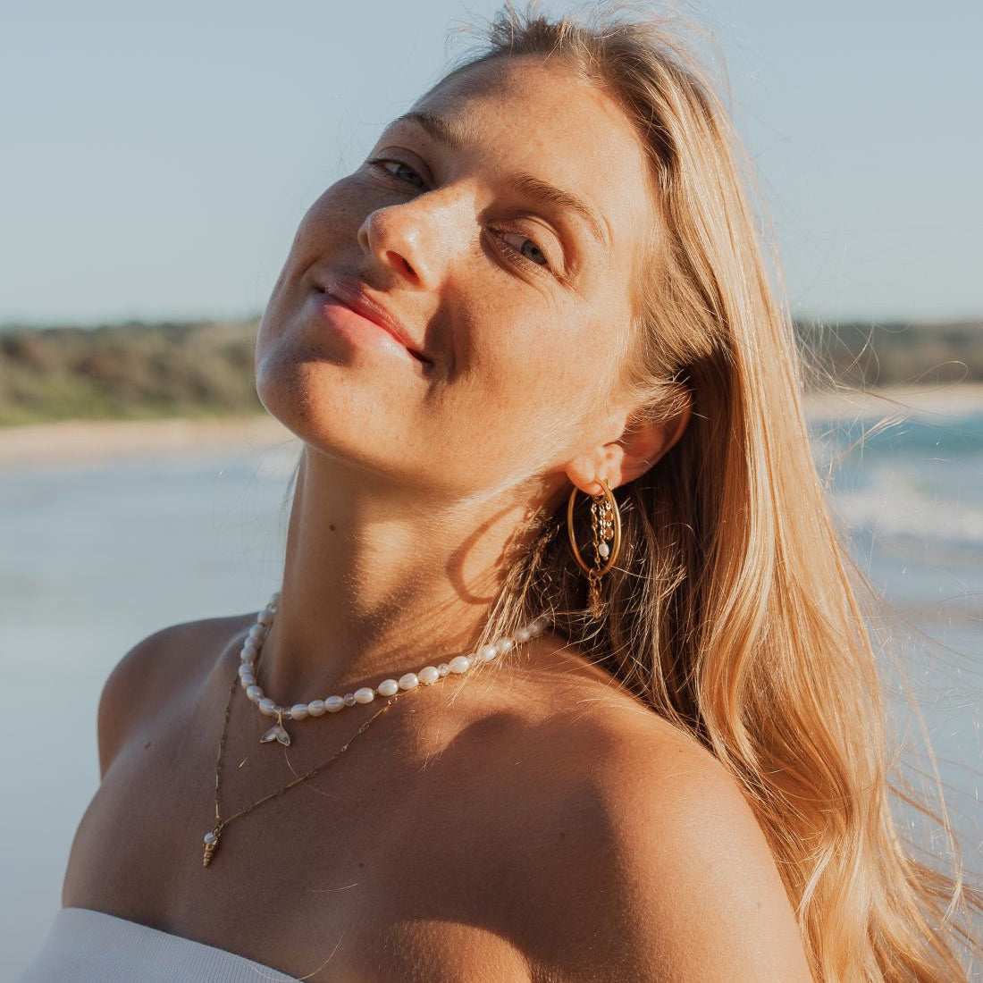 A woman with long blonde hair wearing a white strapless top, a pearl necklace, and gold earrings, posing on a sunny beach with the ocean and sky in the background.
