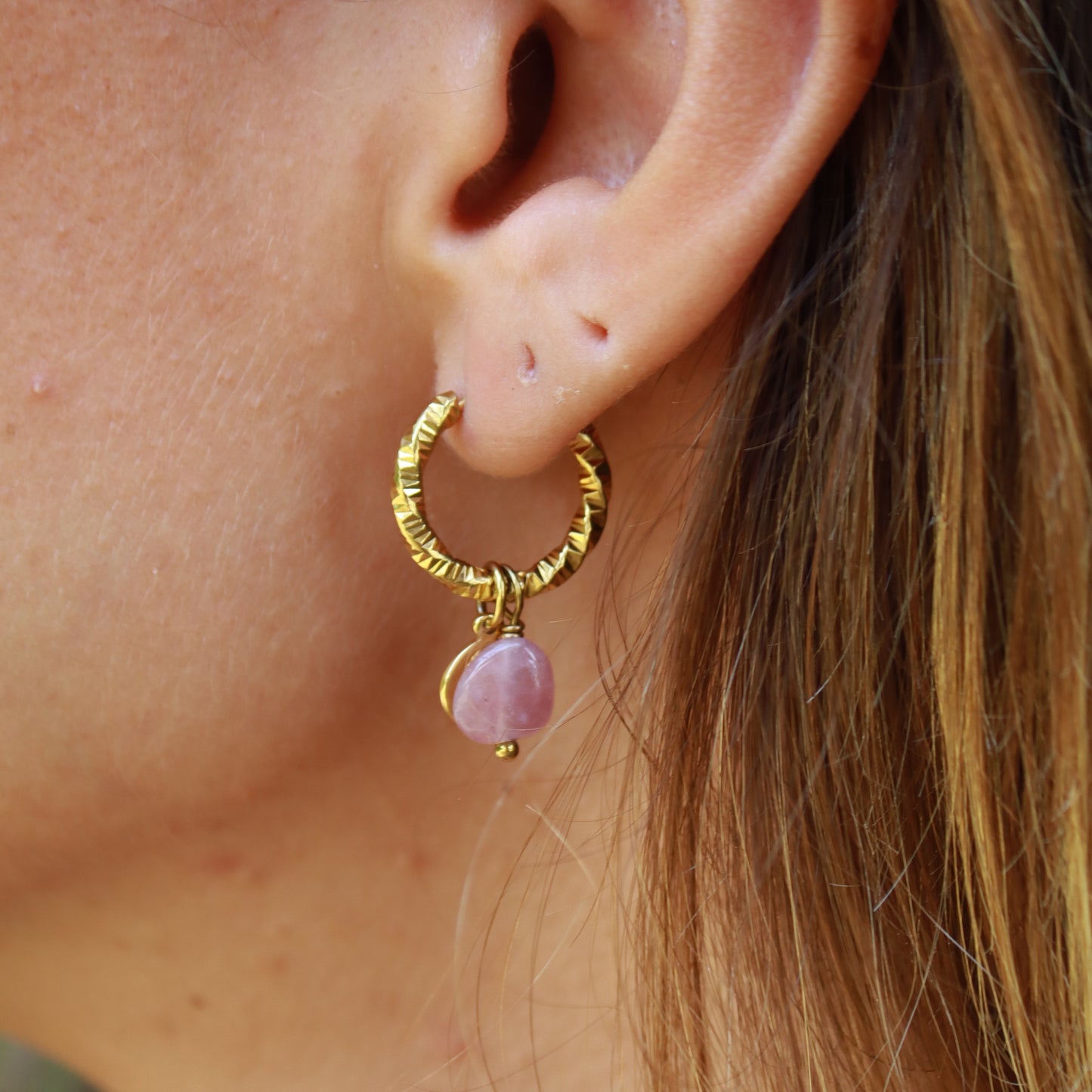 Close-up of an ear with a gold hoop earring featuring a pink gemstone.