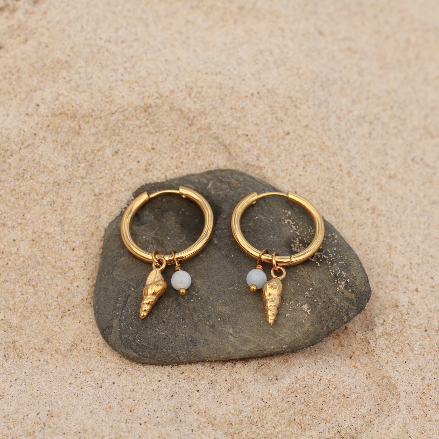 Gold hoop earrings with small charms on a stone in sand
