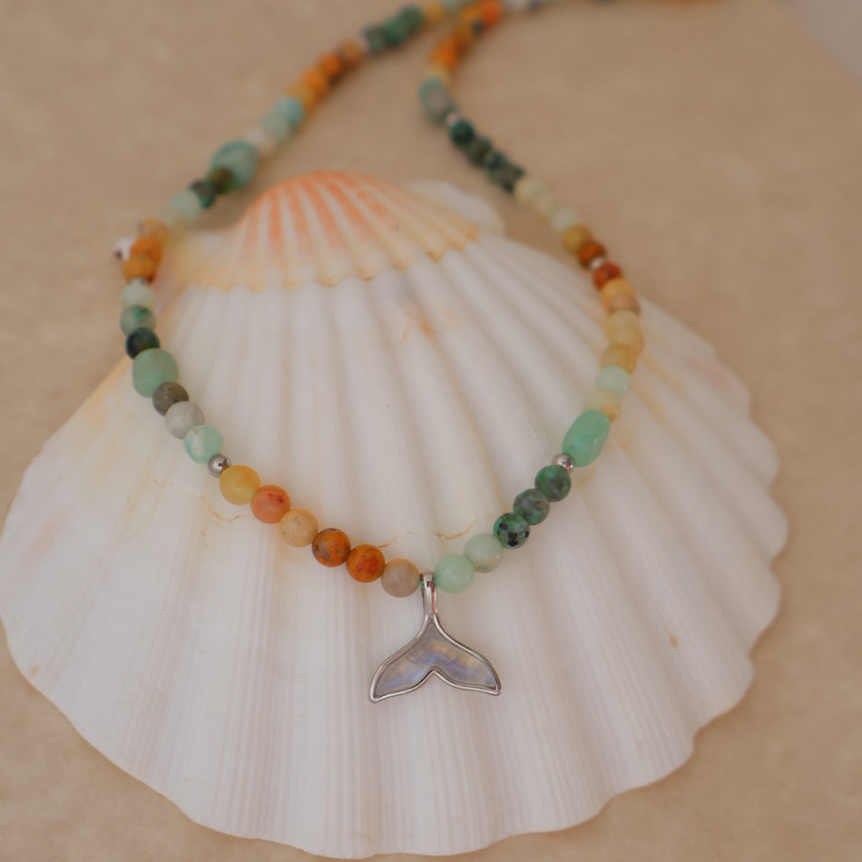 A beaded necklace with a silver whale tail pendant, displayed on a large white seashell. The necklace features small, round beads in various earthy colors including orange, green, yellow, and brown.