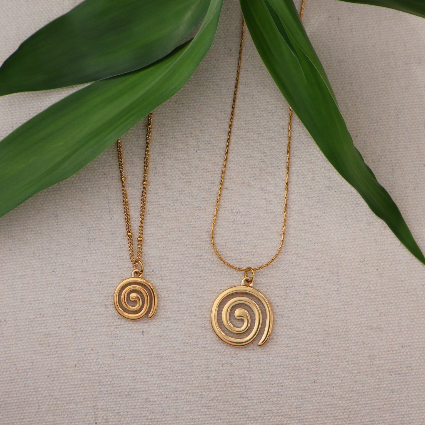 Two gold spiral necklaces on a neutral background with green leaves.
