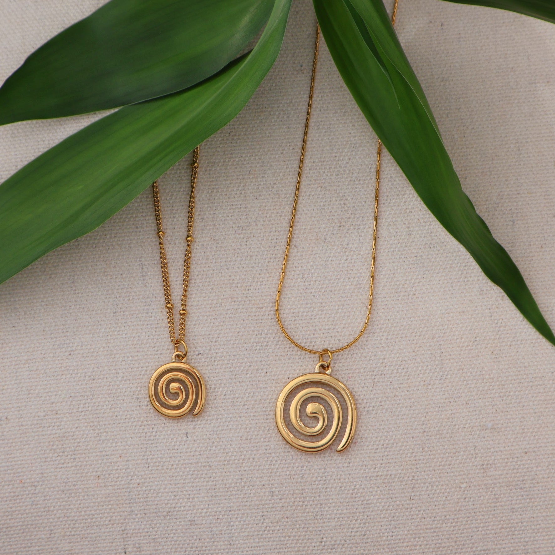 Two gold spiral necklaces on a neutral background with green leaves.