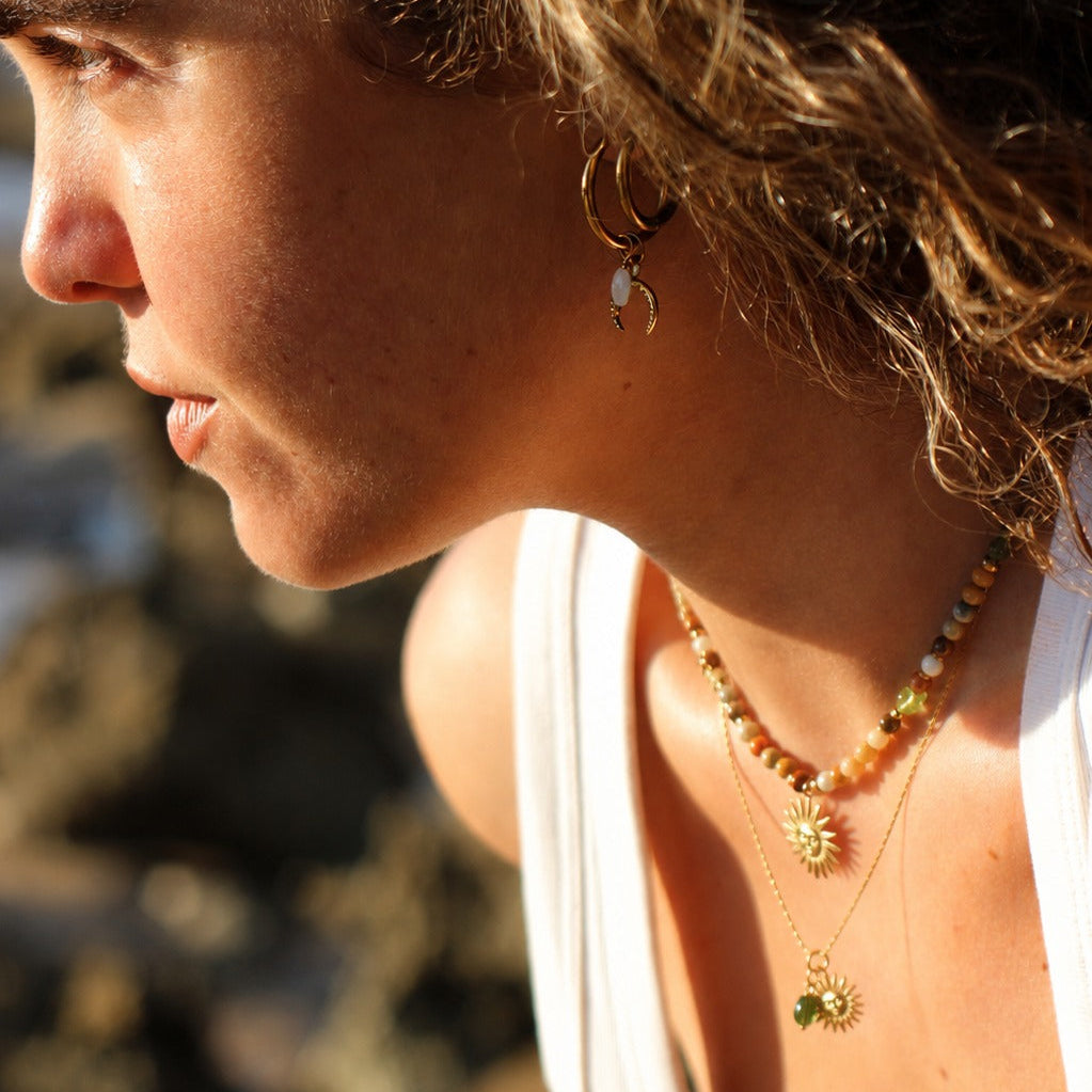A close-up side profile of a woman outdoors wearing delicate gold necklaces, one with a flower-like charm and another layered with green leaf charms, along with a small earring.