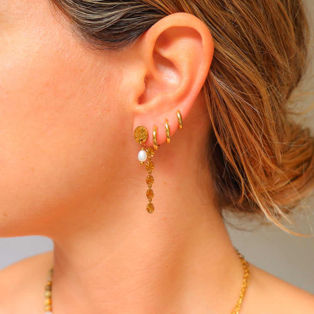 A close-up of a person's ear wearing a stack of gold earrings, including a long dangling earring with a white pearl and faceted gold beads, alongside two small gold hoop earrings.