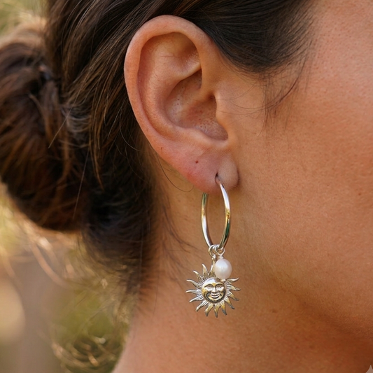 A close-up of a person's ear wearing a silver hoop earring with a dangling sun charm and a small pearl accent.