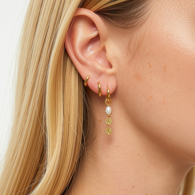 A close-up of a model's ear wearing a stack of gold earrings, including a small hoop, a huggie, and a drop earring with a white pearl and gold leaf details.