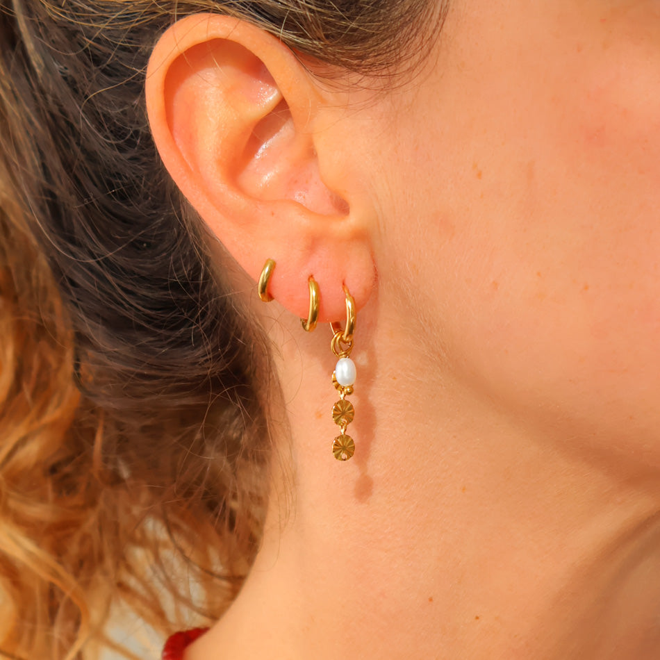 A close-up of a person's ear wearing multiple gold hoop earrings, including a dangling gold hoop with a white pearl and small gold charms.