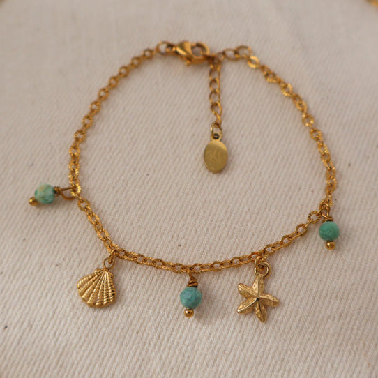 A gold-toned beaded necklace with shell and starfish charms, displayed on a textured off-white fabric background.