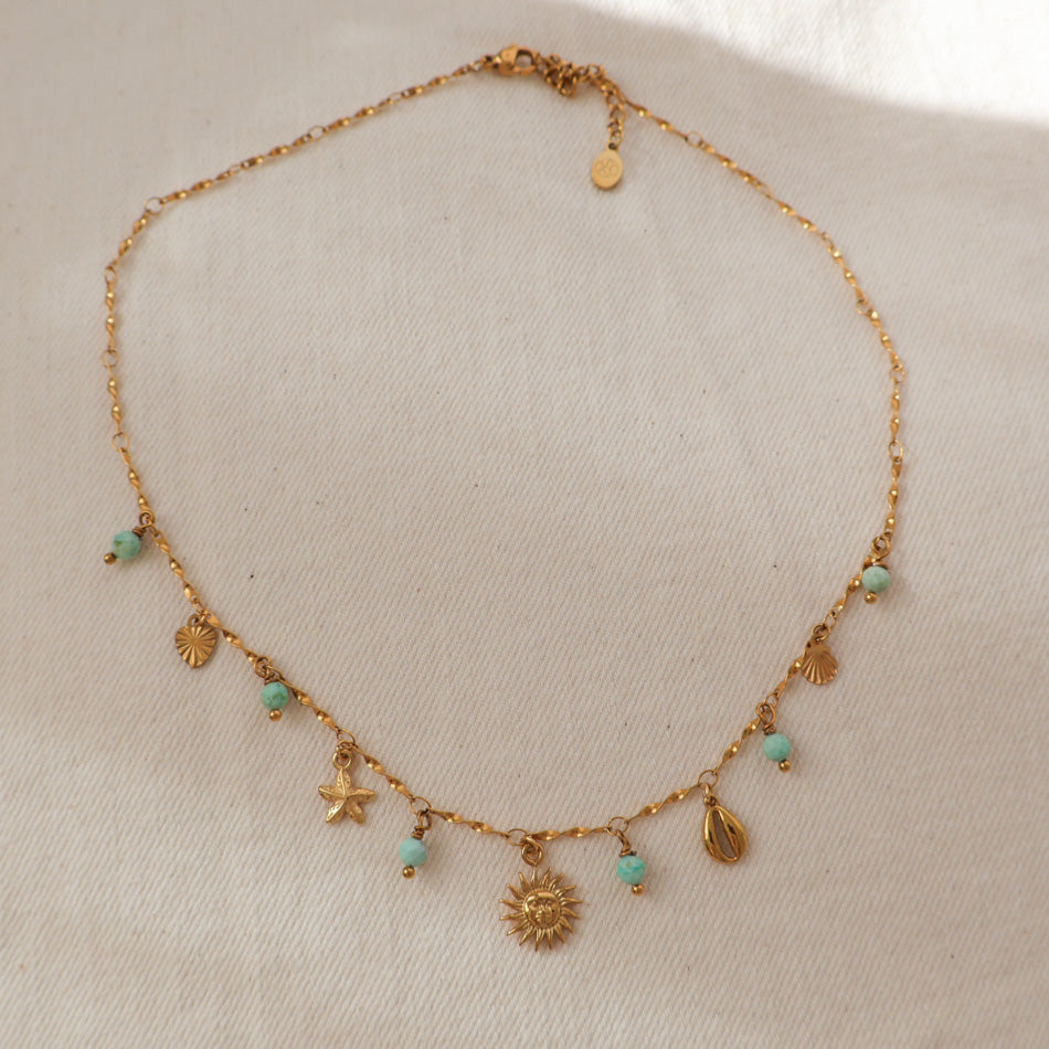 A gold chain necklace with small dangling charms, including a sun, starfish, shells, and turquoise-colored beads, displayed on a textured off-white fabric background.