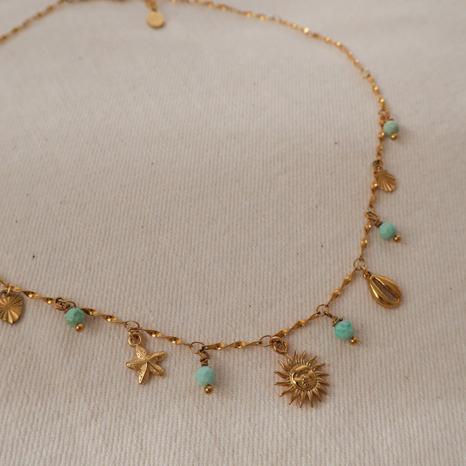 A delicate gold-tone chain necklace with small sea-themed charms including a sun, starfish, seashell, and star, accented by small turquoise beads, displayed on a textured off-white fabric background.