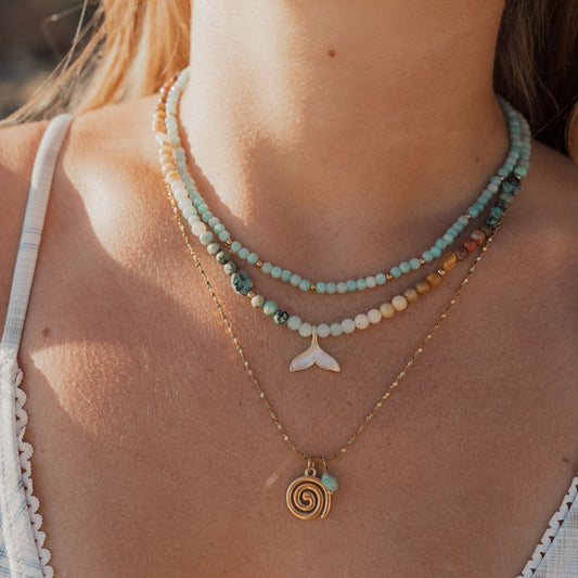 A close-up lifestyle shot of a woman wearing a layered necklace set. The set includes a multi-strand beaded necklace with light blue, white, and gold beads, a gold chain with a white shell-shaped pendant, and a gold chain with a spiral shell pendant.