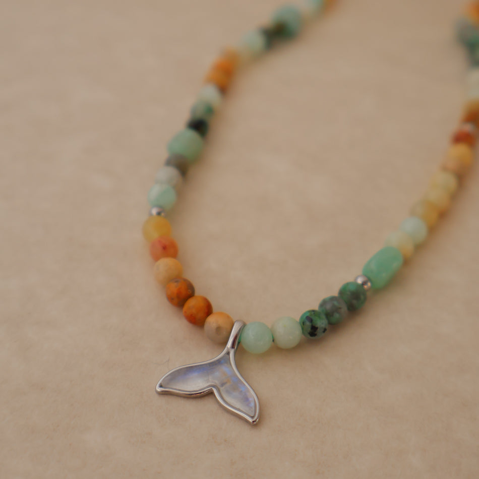 A beaded necklace with a silver whale tail pendant, featuring a mix of green, orange, yellow, and brown beads on a beige background.