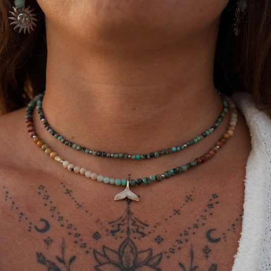 Close-up of a person's neck and upper chest wearing two beaded necklaces. The top necklace has turquoise and brown beads with a silver whale tail pendant, and the bottom necklace has small white and turquoise beads.
