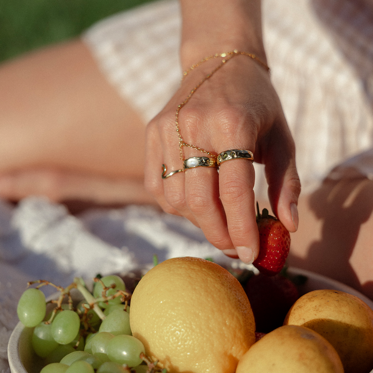 A close-up lifestyle shot of a person's hand wearing gold rings, reaching for a strawberry over a plate of fruit (grapes, lemon, pears) on a white checkered blanket outdoors.