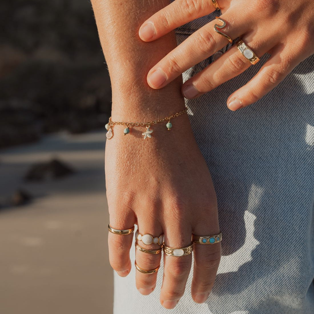 A close-up lifestyle shot of a person's hands and arms adorned with multiple delicate gold jewelry pieces, including rings, bracelets, and a chain necklace, set against a softly blurred beach background.